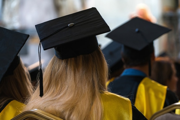 Students wearing gowns and hats sitting indoors, waiting to receive diplomas.