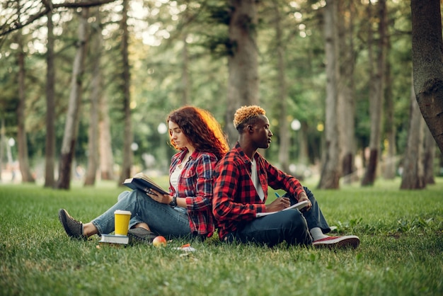 Photo students with book sitting on the grass with their backs to each other, summer park. male and female teenagers studying outdoors and having lunch