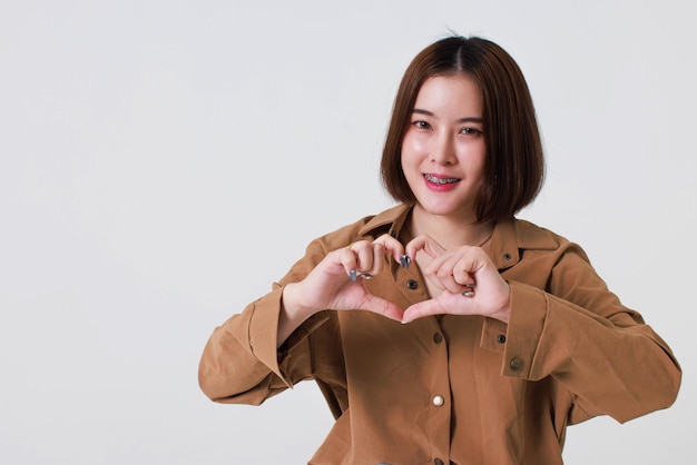 Studio cutout shot of Asian young pretty short hair female model in long brown coat jacket stand smile crossed arm hold thumb up pointing presenting product on blank copy space on white background.