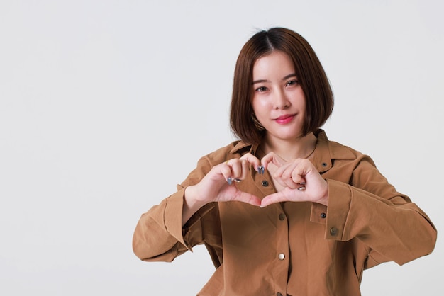 Studio cutout shot of Asian young pretty short hair female model in long brown coat jacket stand smile crossed arm hold thumb up pointing presenting product on blank copy space on white background.