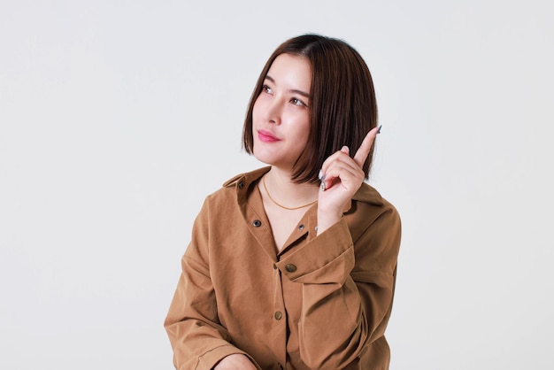 Studio cutout shot of Asian young pretty short hair female model in long brown coat jacket stand smile crossed arm hold thumb up pointing presenting product on blank copy space on white background.