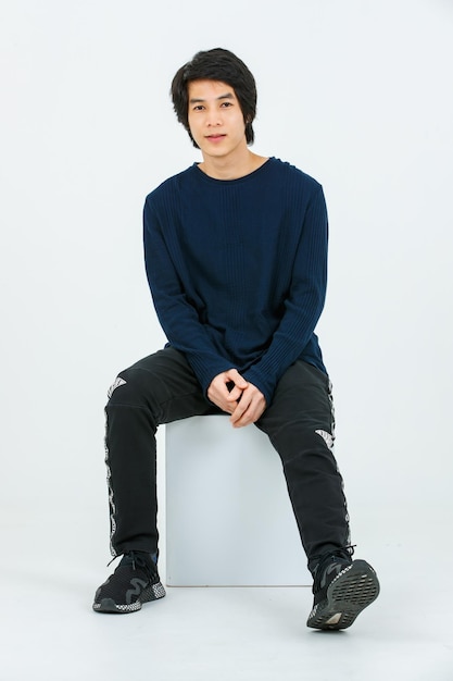 Studio full body shot of Asian young handsome confident slim healthy athletic teenager fashion male model in long sleeve shirt fabric pants and sneaker sitting on box posing on white background