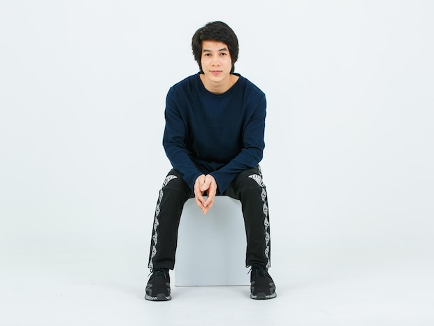 Studio full body shot of Asian young handsome confident slim healthy athletic teenager fashion male model in long sleeve shirt fabric pants and sneaker sitting on box posing on white background