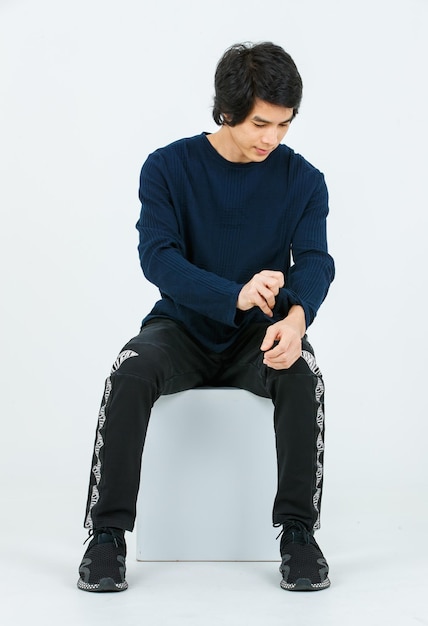 Studio full body shot of Asian young handsome confident slim healthy athletic teenager fashion male model in long sleeve shirt fabric pants and sneaker sitting on box posing on white background