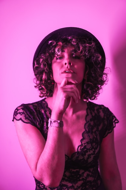 Studio lifestyle, seductive portrait of a young Caucasian woman in a beautiful outfit and a hat, illuminated with a pink neon light