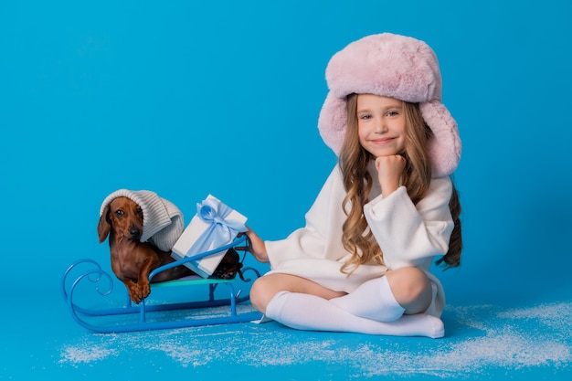 studio photo of a girl in a white sweater and hat and a dachshund dog sitting in a sled