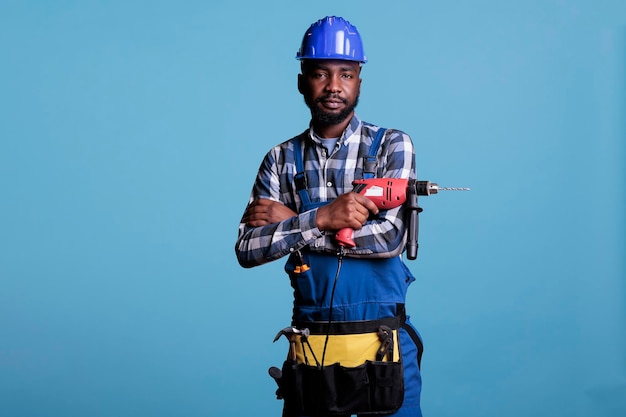 Studio photo of repairman looking at the camera, arms crossed over his chest holding cordless electric drill. Construction worker wearing overalls and belt with all working tools.