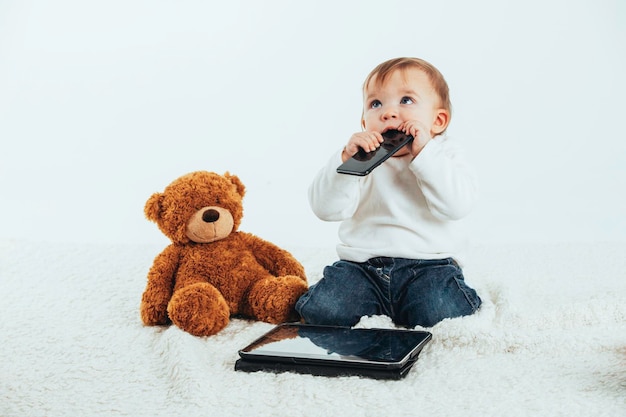 Studio photo with the white background of a baby39s face with a mobile in his mouth next to a teddy bear and a tablet