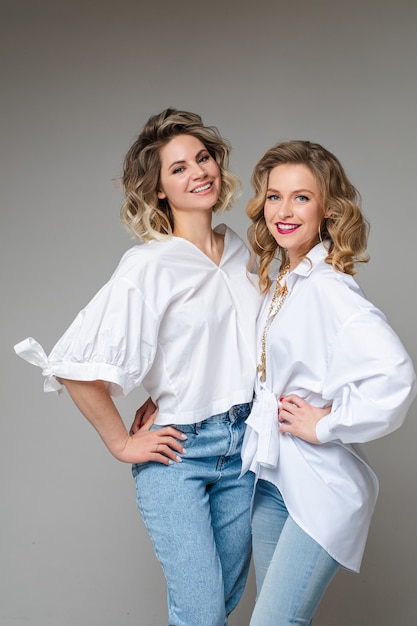 Studio portrait of attractive blonde adult women in white top and jeans posing snuggling on grey wall