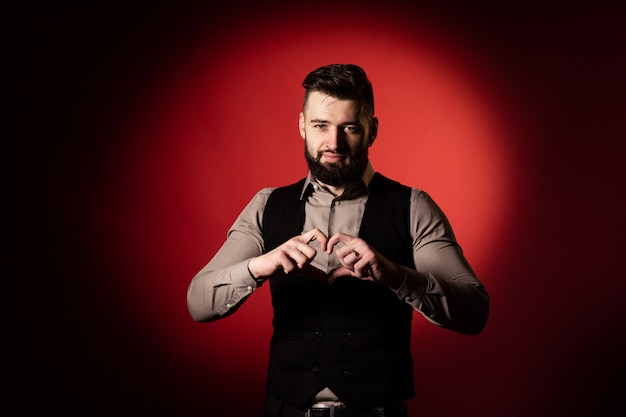 Studio portrait of a bearded man in a vest on a red background. The man folded his fingers in the form of a heart