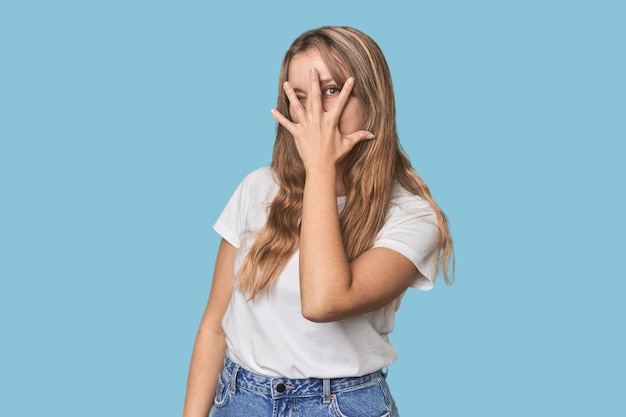 Studio portrait of a blonde Caucasian woman blink at the camera through fingers embarrassed