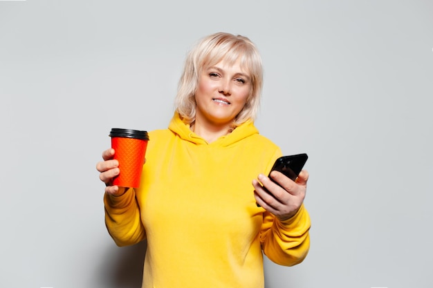 Studio portrait of blonde woman using smartphone holding paper cup for coffee takeaway of red color wearing yellow sweater on white background