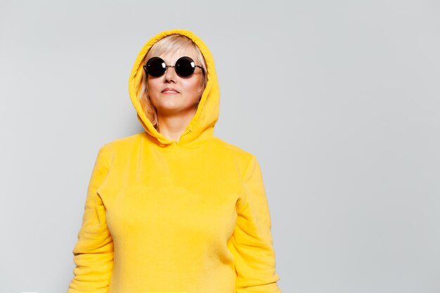 Studio portrait of blonde woman in yellow hoodie sweater wearing round sunglasses on white background with copy space