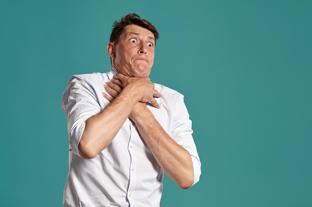 Studio portrait of a brunet young businessman in a classic white shirt choking himself while posing over a blue background. Stylish haircut. Sincere emotions concept. Copy space.