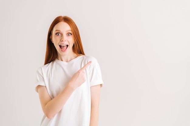 Studio portrait of cheerful attractive young woman with open mouth recommending suggest select advert point index finger aside on isolated white background, commercial promo area, mock up copy space.