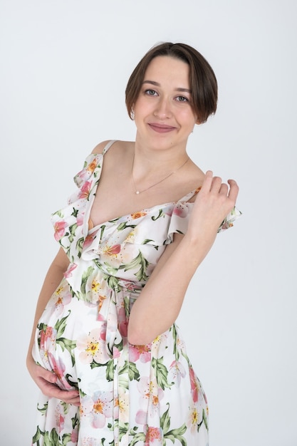 Studio portrait of cheerful young pregnant woman in summer dress on white gray wall, happy pregnancy concept