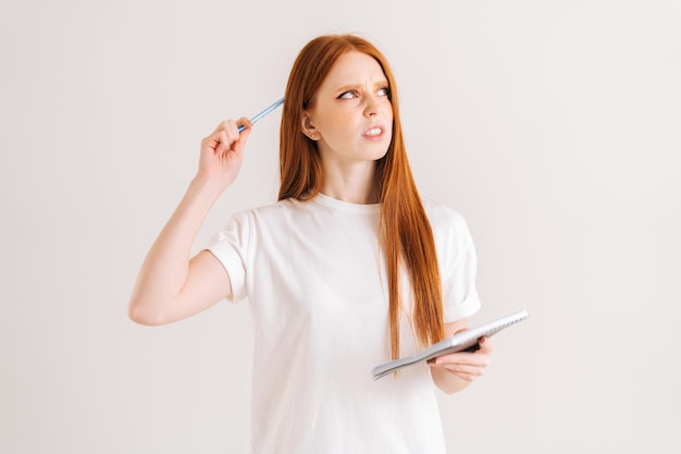 Studio portrait of confused redhead young woman scratches head with pencil holding diary looking