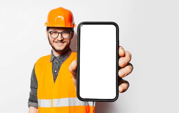 Studio portrait of construction worker holding smartphone with blank screen on white background Mockup concept Happy young man Orange safety equipment for engineers