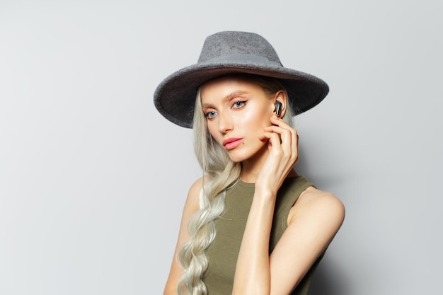 Studio portrait of cute blonde girl touching wireless earphones while listening to music on white background Wearing grey hat