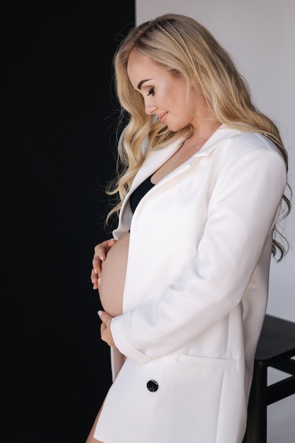 Studio portrait of gogreous pregnant woman in white jacket hold her hand on belly and looks down