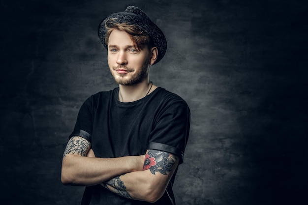 Studio portrait of handsome male with crossed tattooed arms, dressed in a black t shirt and tweed hat.