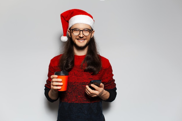 Studio portrait of handsome smiling guy holding paper cup with drink takeaway and smartphone in hands Wearing Santa Claus hat Christmas concept White background