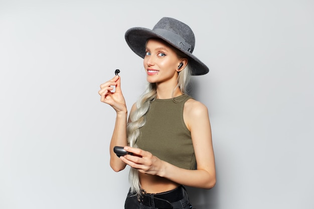 Studio portrait of happy pretty blonde girl holding wireless earphones and charger case in hands wearing grey hat on white background