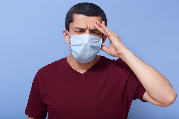 Studio portrait of ill brunette with frowned facial expression, having headache, closing eyes, being in medical mask, putting fingers at forehead, having fever, being infected, feeling weakness.