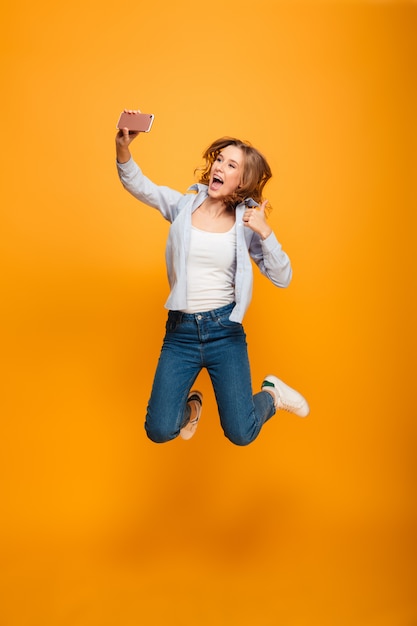 Studio portrait of joyful woman wearing jeans and sneakers smiling and showing thumb up while taking selfie with cell phone, isolated over yellow space