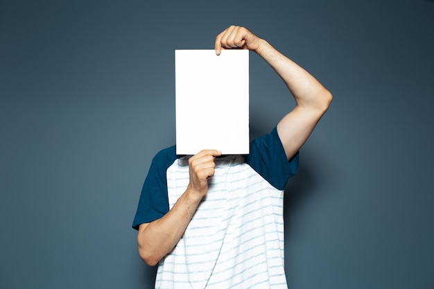 Studio portrait of man holding empty vertical paper board on face Copy space Blue background