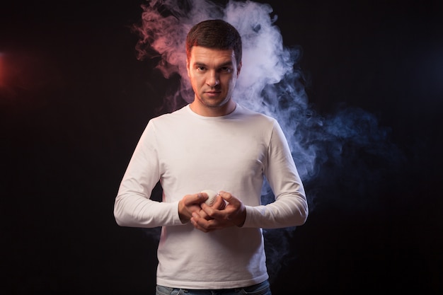 Studio portrait of muscular sportsman player posing on black background in colored smoke with a baseball in his hand. 