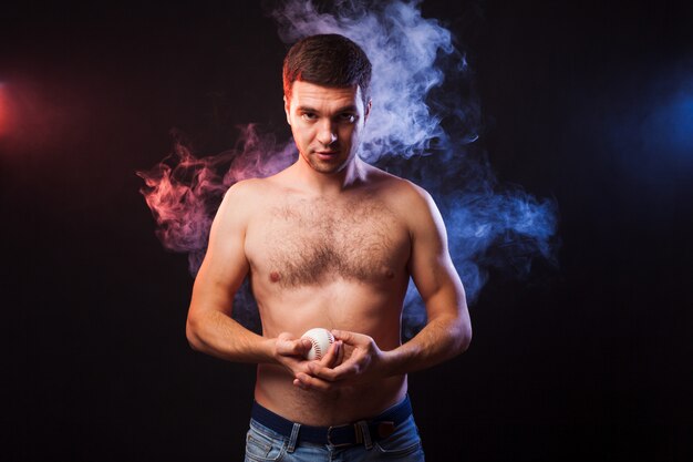 Studio portrait of muscular sportsman with player posing on black background in colored smoke with baseball in his hand. 