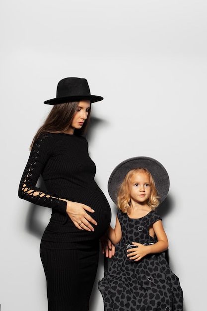 Studio portrait of pregnant smiling woman and child girl wearing hats on white background Mother and daughter in black