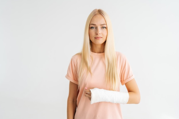 Photo studio portrait of pretty injured blonde young woman with broken forearm wrapped in plaster bandage looking at camera standing on white isolated background