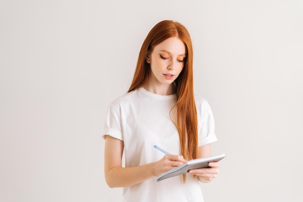 Studio portrait of pretty redhead young woman writing in notebook looking down standing on white isolated background Confident female student making notes in paper diary