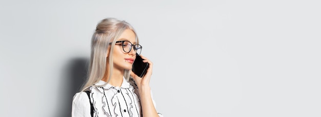 Studio portrait of pretty smiling girl with blonde hair talking on smartphone wearing eyeglasses on white background panoramic banner view with copy space