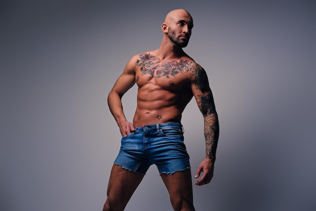 Studio portrait of shirtless shaved head, muscular male with tattoos on his torso dressed in a denim shorts over grey vignette background.