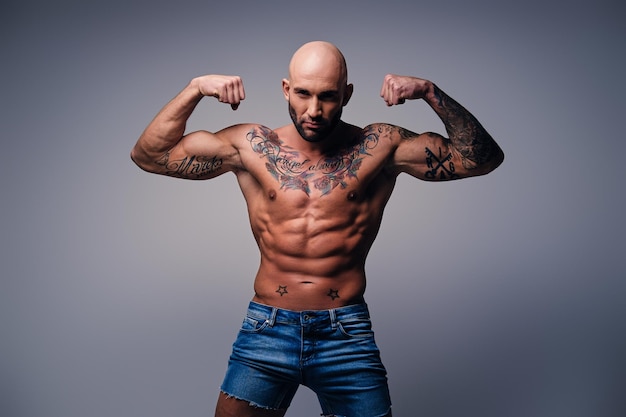 Studio portrait of shirtless shaved head, muscular male with tattoos on his torso over grey vignette background.