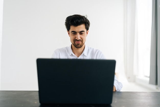 Studio portrait of smiling bearded man in shirt working typing on laptop computer looking to screen sitting at desk in room with light interior