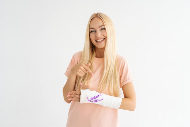 Studio portrait of smiling blonde young woman with broken arm wrapped in plaster bandage with funny inscription looking at camera