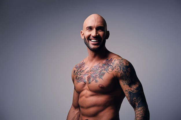 Studio portrait of smiling shirtless athletic shaved head male with tattoos on his torso posing over grey vignette background.