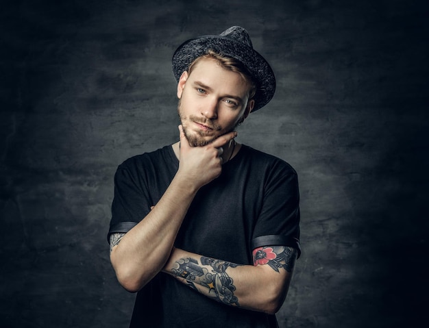 Studio portrait of thoughtful male with crossed tattooed arms, dressed in a black t shirt and tweed hat.