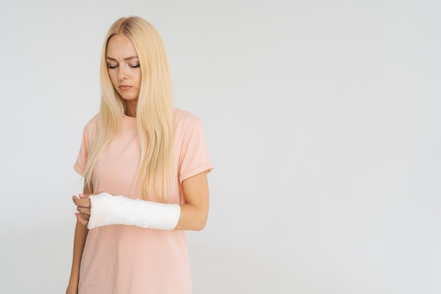 Studio portrait of unhappy injured blonde young woman with broken arm wrapped in plaster bandage looking down standing on white isolated background