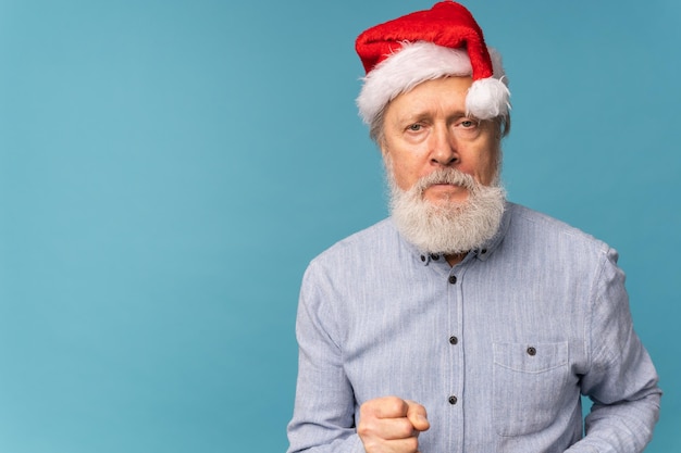 Studio portrait of white beard old man in Santa hat showing fist and looking at camera with angry expression copy space emotion and bad christmas mood concept