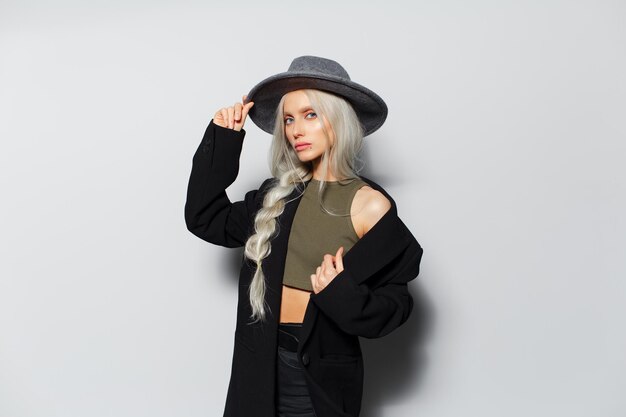 Studio portrait of young beautiful blonde girl with a braid wearing black coat keeps hand on hat on white background