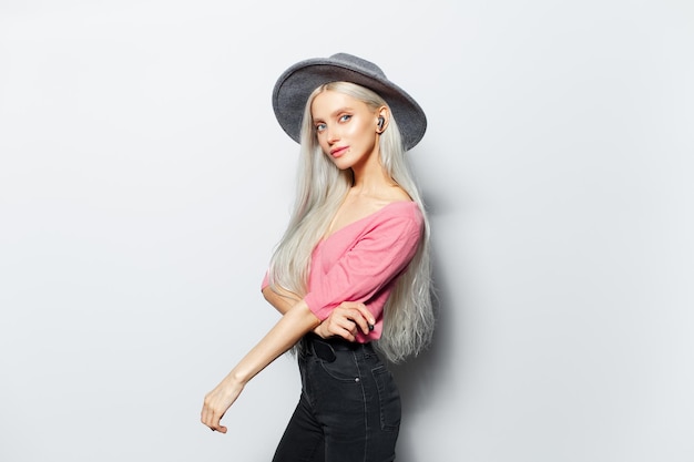 Studio portrait of young beautiful blonde girl with wireless earphones in ears wearing grey hat and pink shirt on white background