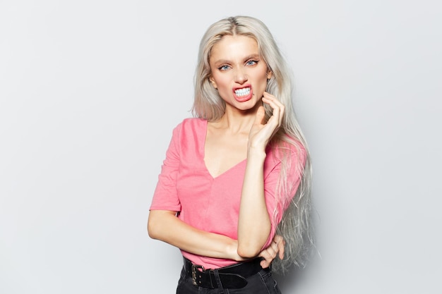 Studio portrait of young blonde pretty girl showing teeth wearing pastel pink shirt on white background