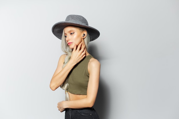 Studio portrait of young blonde pretty girl with earbuds in ears on white background wearing grey hat