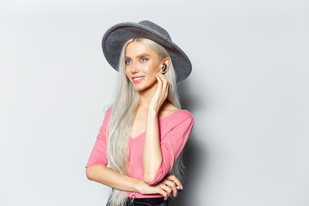 Studio portrait of young blonde smiling girl listening the music via earbuds on white background wearing grey hat and pink shirt