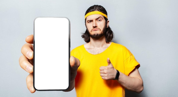 Studio portrait of young confident man holding big smartphone with blank on screen in hand showing close to camera a device with mockup and thumb up on white Wearing yellow shirt and head band